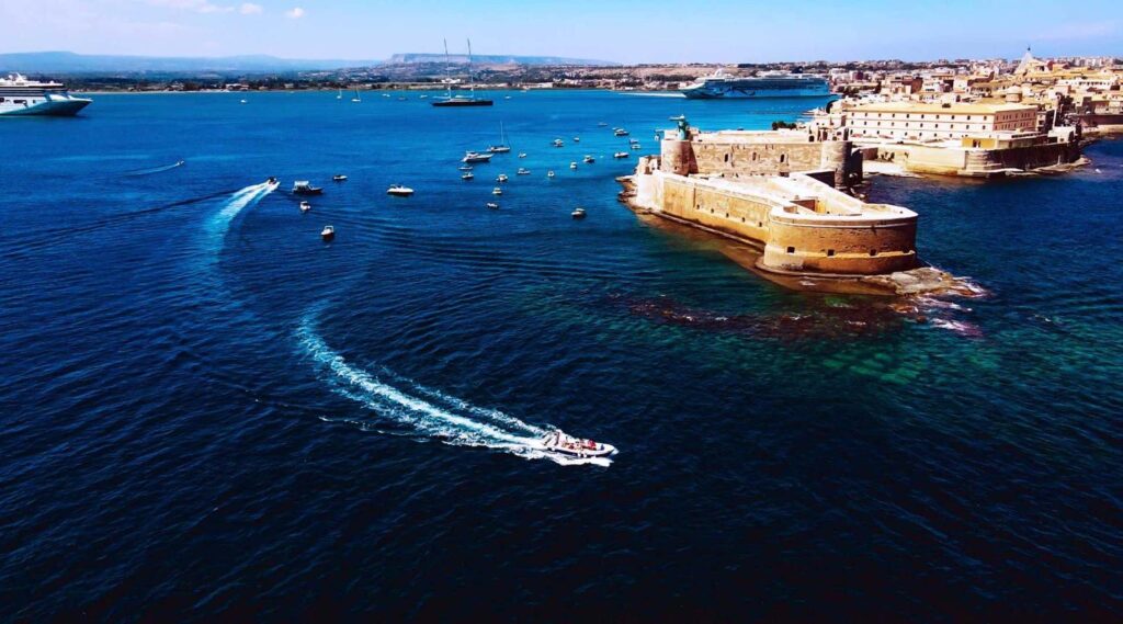 Boat cruising along the historic coastline of Syracuse in Sicily with limestone cliffs and sea caves.