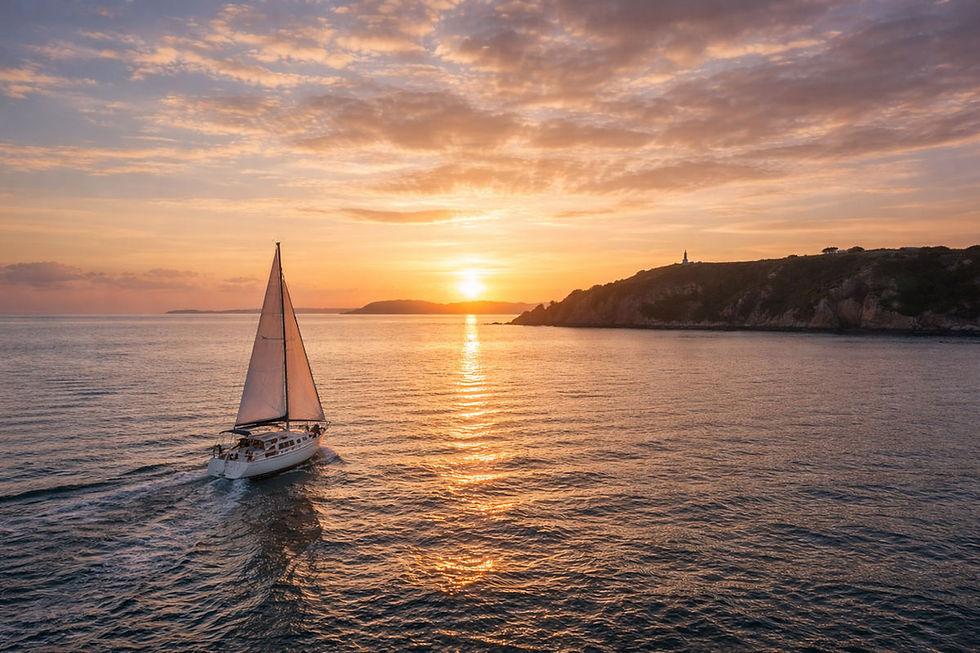 Sailboat cruising past the Cornish coastline at sunset with a lighthouse in the distance.